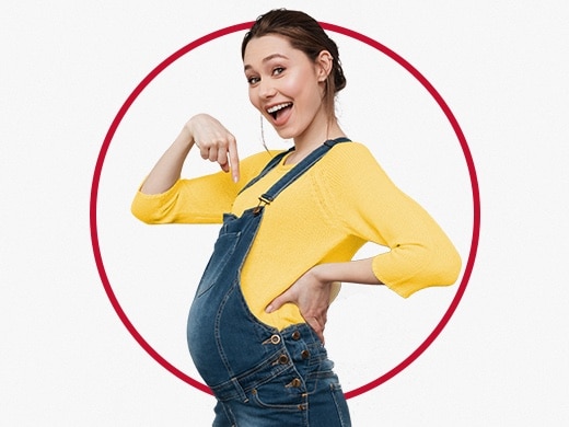 Vesela mlada noseča ženska v rumenem puloverju kaže na svoj trebušček